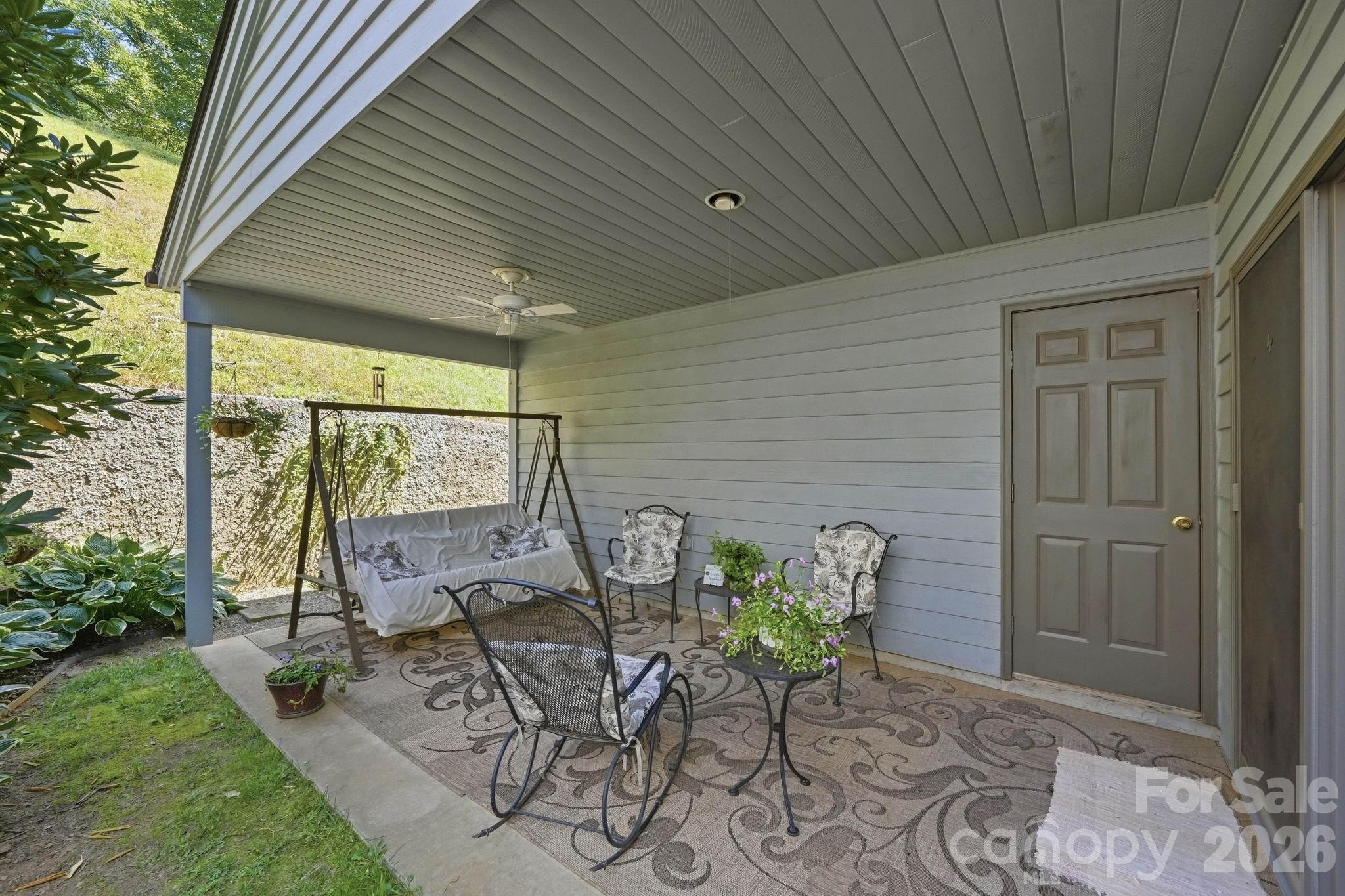 Covered patio with a cushioned swing, seating, ceiling fan, and potted flowers.