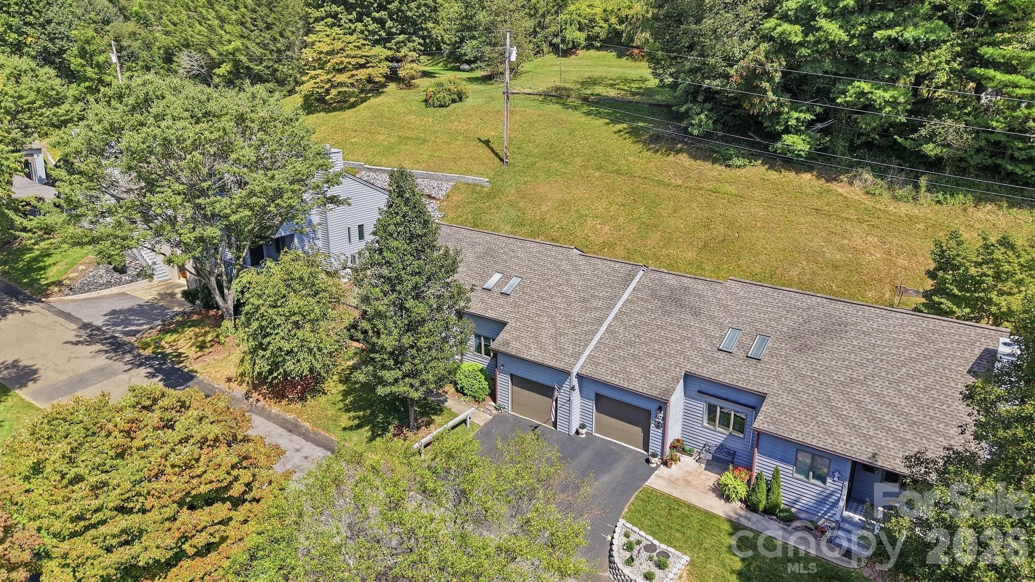 Aerial view of blue-gray attached homes with garages beside a broad grassy area and wooded edge.