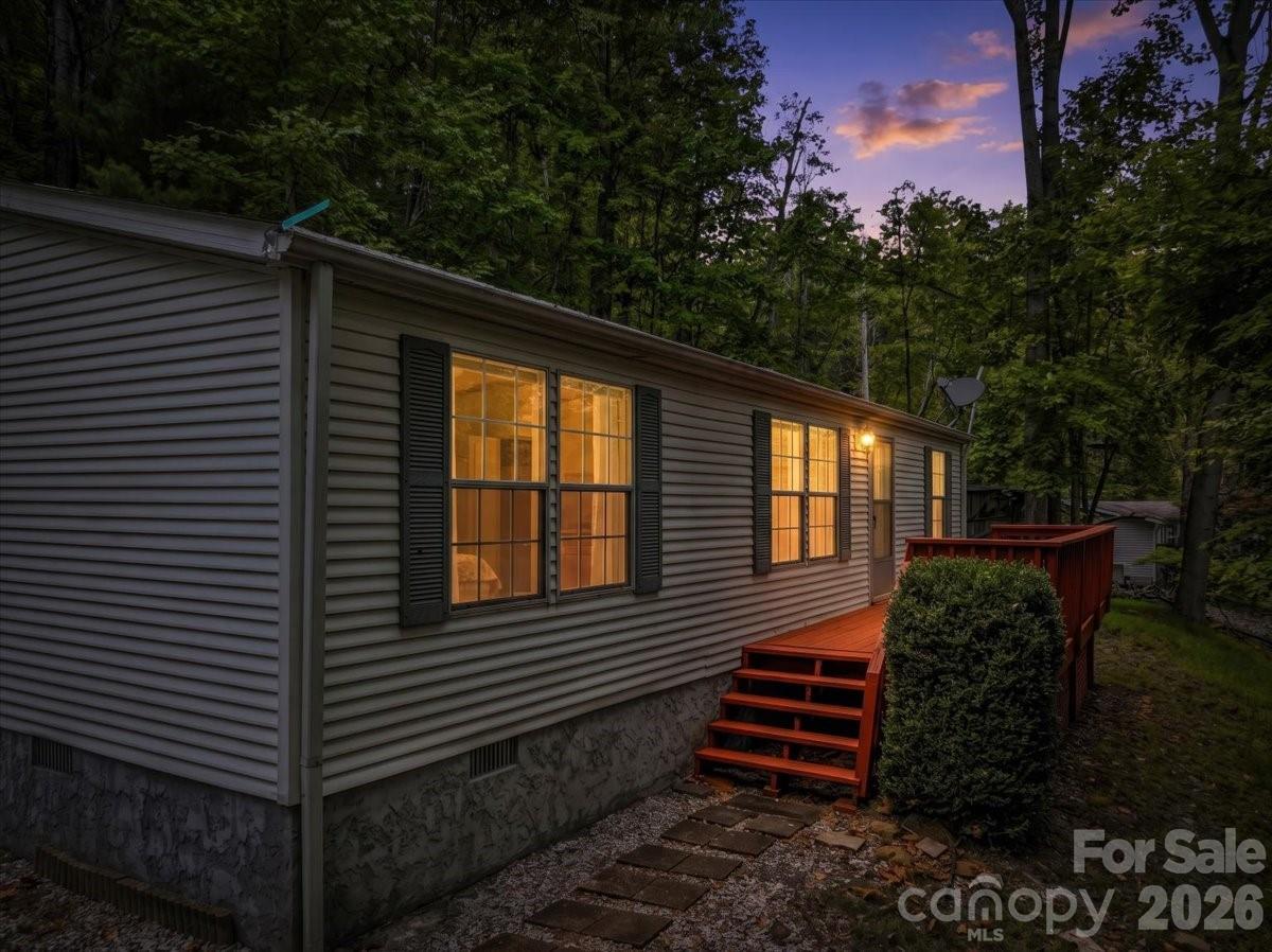 Home exterior at dusk with warmly lit windows, red deck steps, and wooded surroundings.