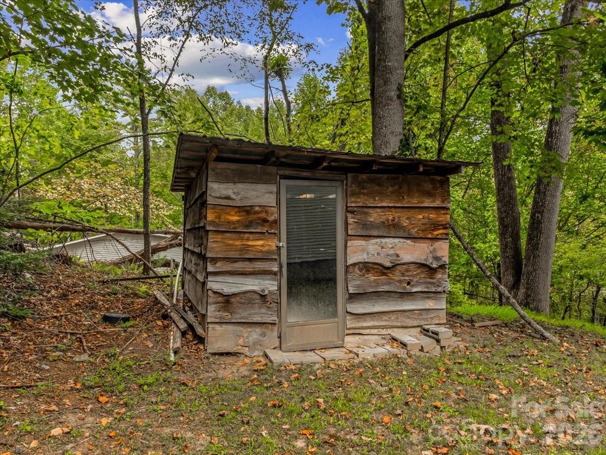 Weathered wooden outbuilding with a screened door beneath a metal roof in a leafy wooded setting.