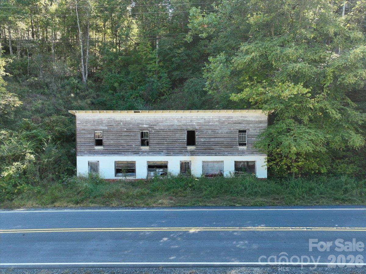 Two-story building with weathered wood siding beside a wooded hillside and roadside.