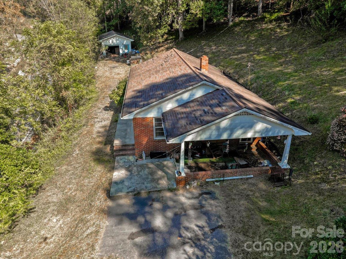 Aerial view of a brick house with a covered porch, driveway, and small outbuilding on a wooded lot.