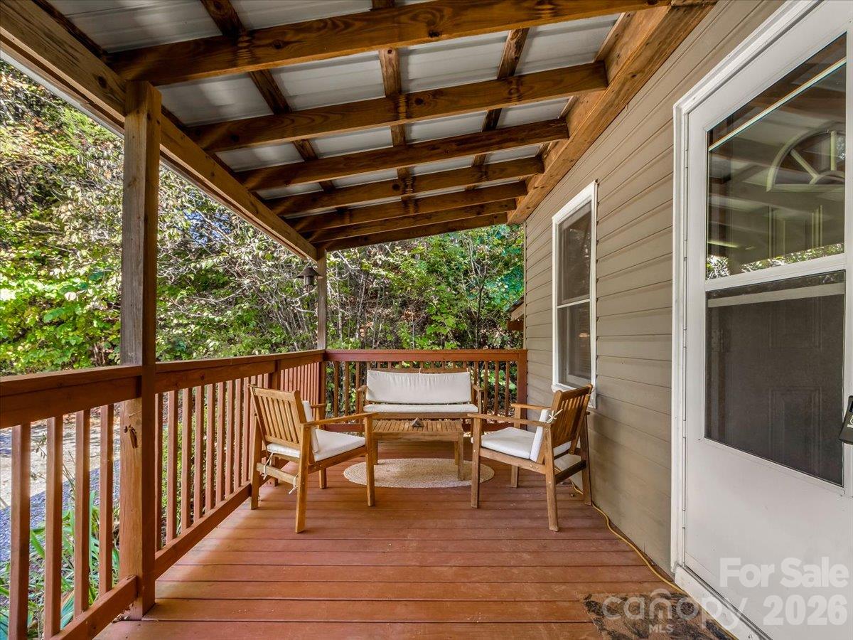 Covered wooden porch with exposed beams and cushioned seating overlooking leafy trees.