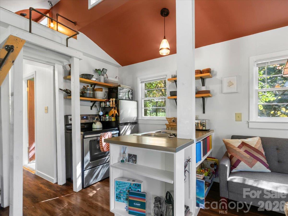 Open-plan kitchen with stainless steel appliances, a sink peninsula, and a gray sofa beneath a window.