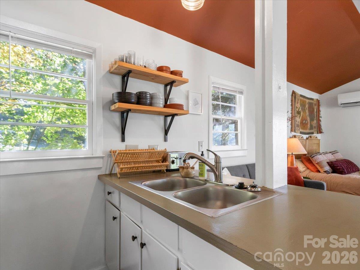 Kitchen with a double-basin sink, open wooden shelves, and large windows overlooking leafy trees.