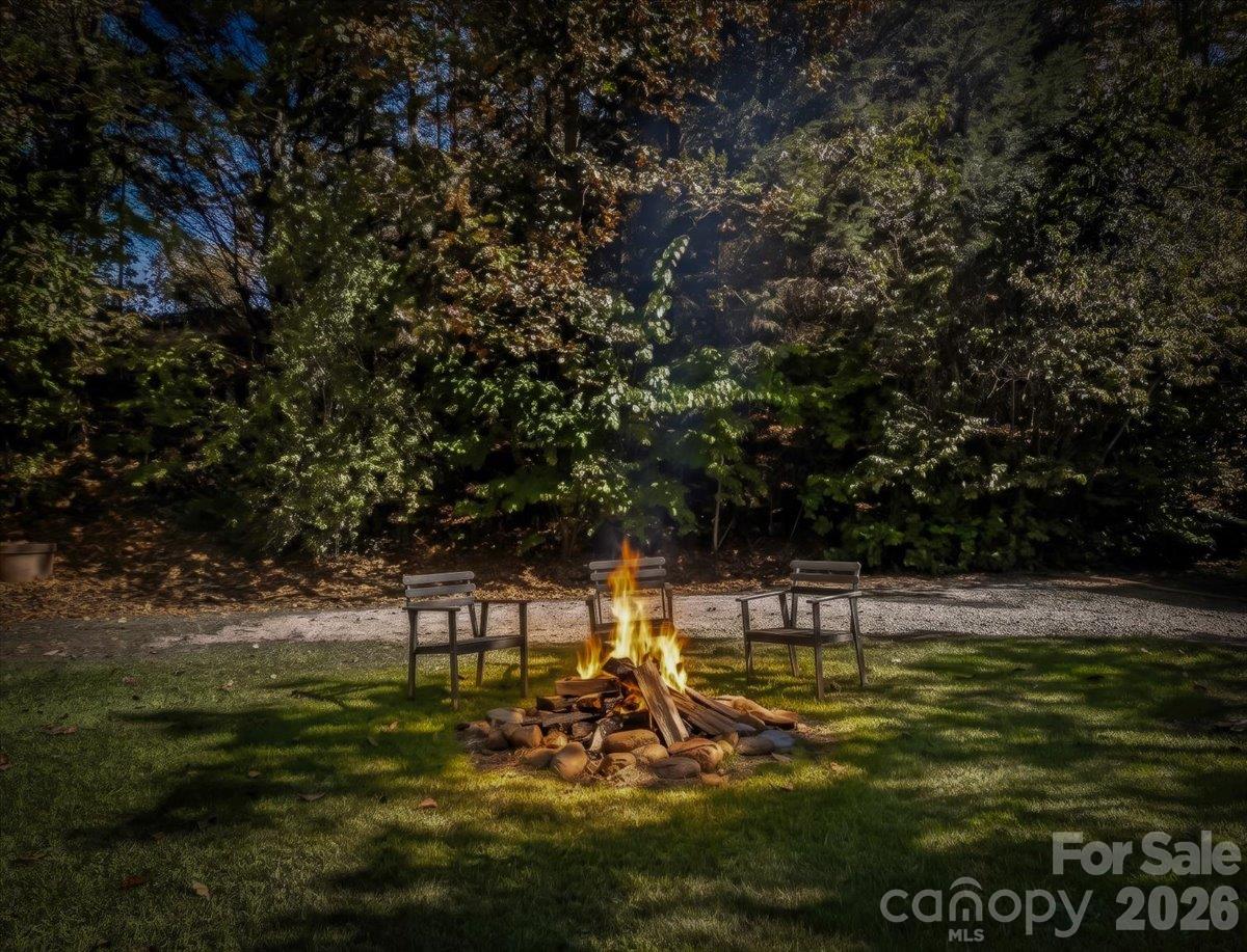 Backyard fire pit surrounded by chairs, with a gravel path and wooded trees behind it.