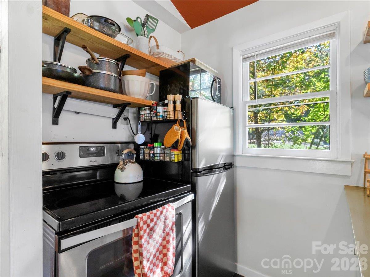 Compact kitchen with stainless steel appliances, open wooden shelves, and a large sunlit window.