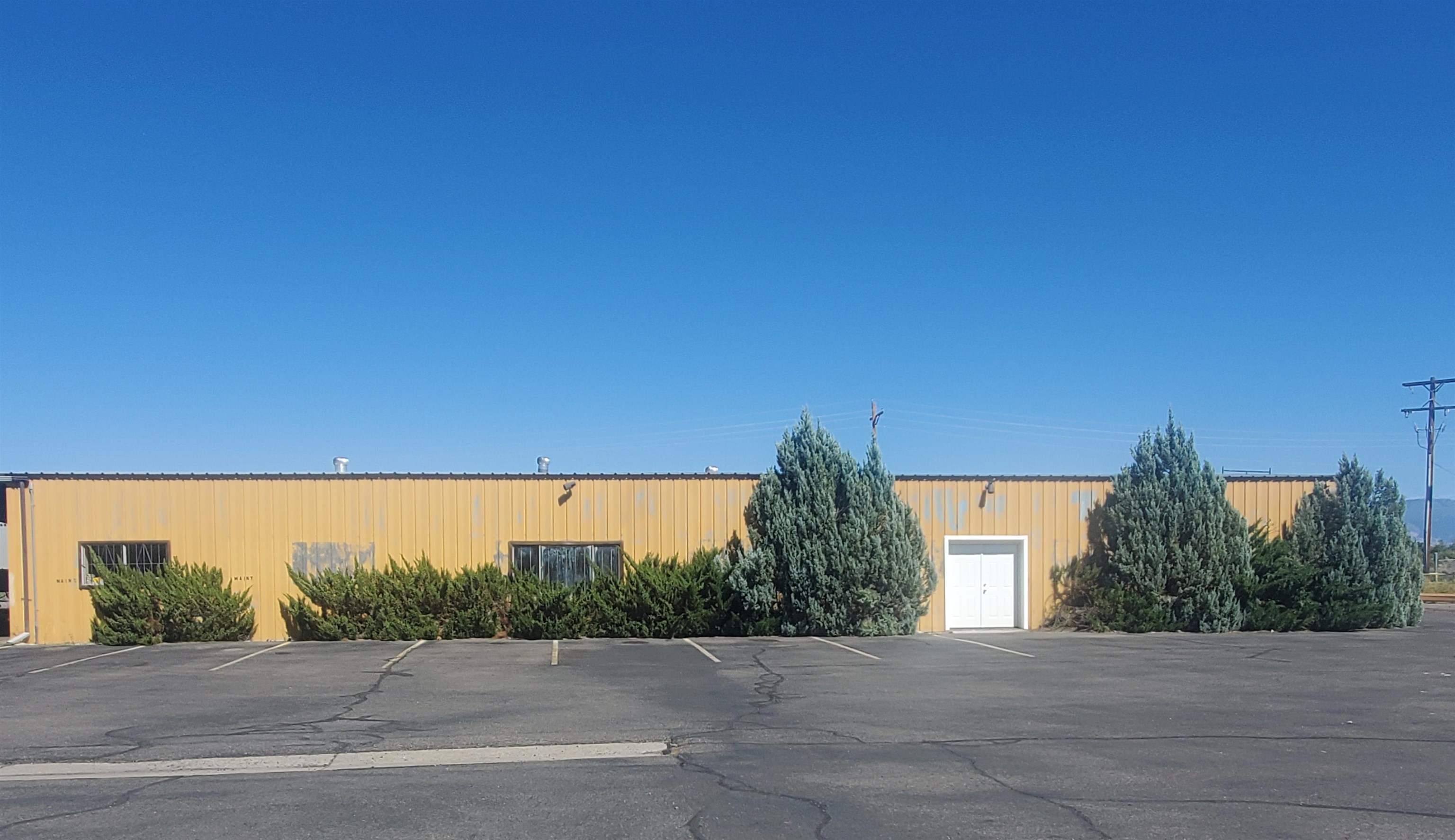 Yellow metal warehouse with a white double door, shrubs, and a cracked asphalt parking lot.