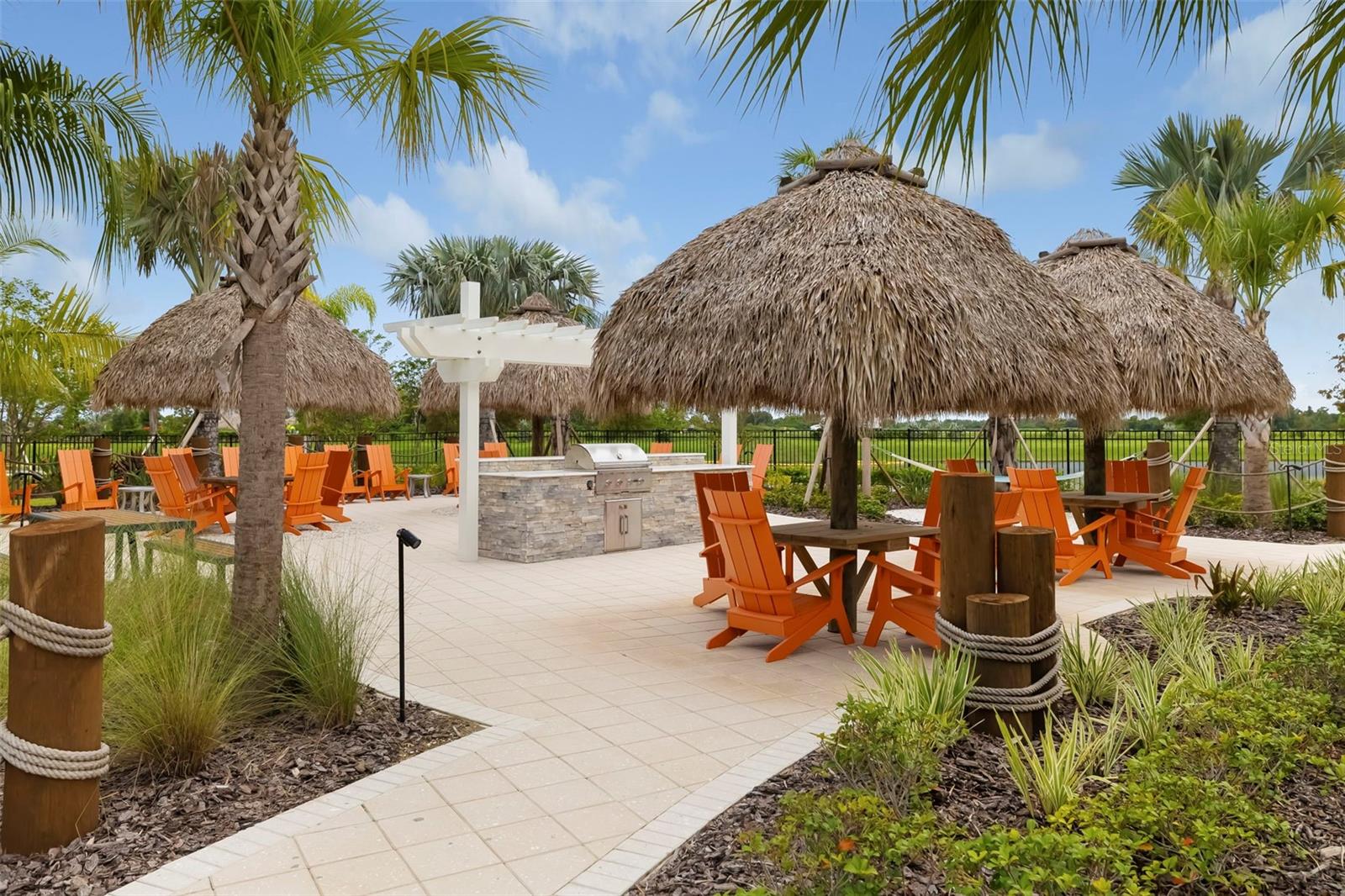 Outdoor gathering area with thatched tiki shelters, orange chairs, and a stone grill.