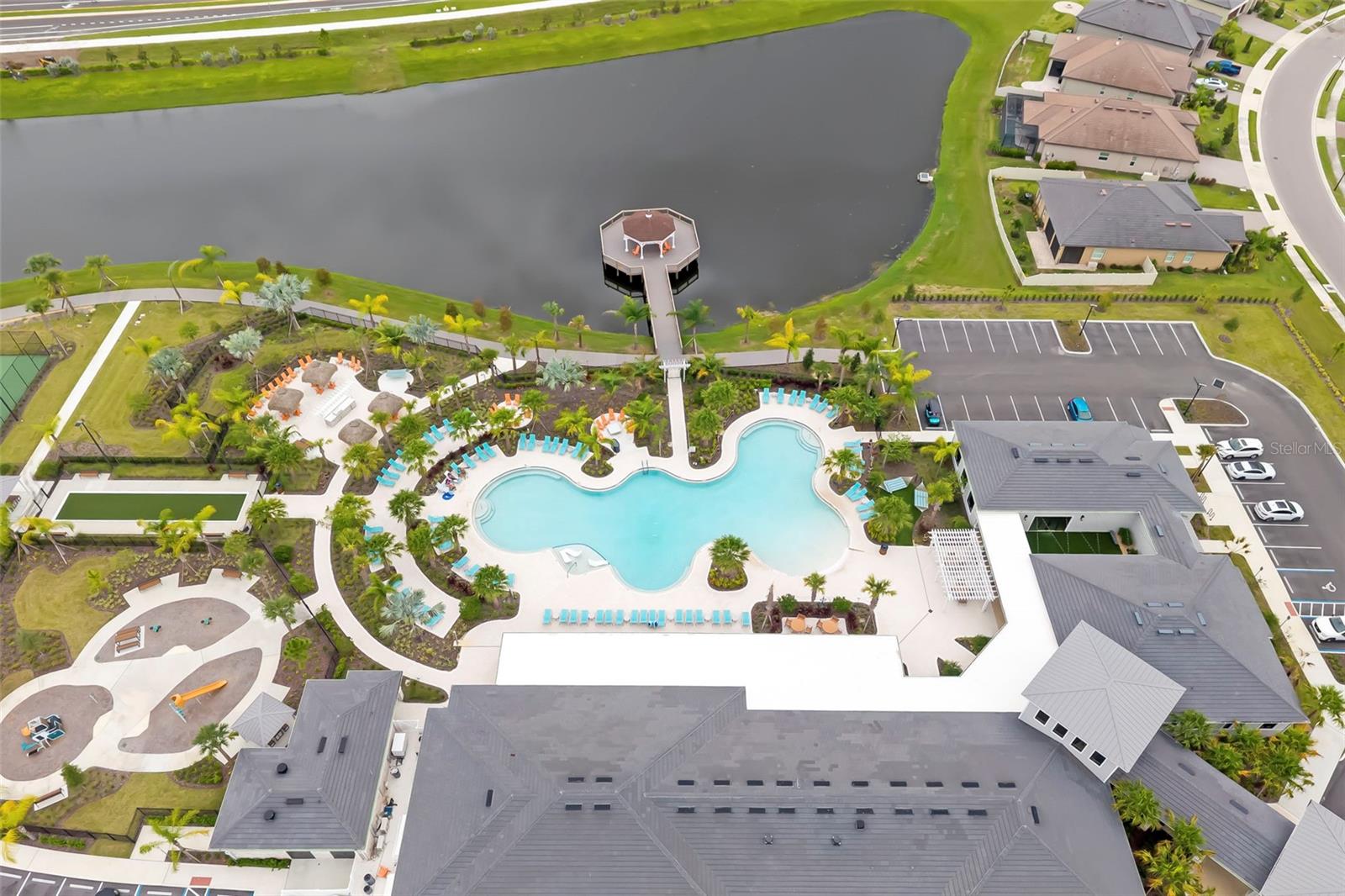 Aerial view of a community pool beside a lake, with a dock, gazebo, and clubhouse.