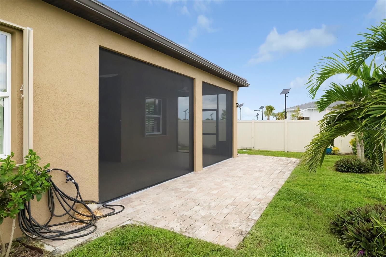 Home’s rear patio with broad pavers, screened openings, and a fenced lawn with palms.