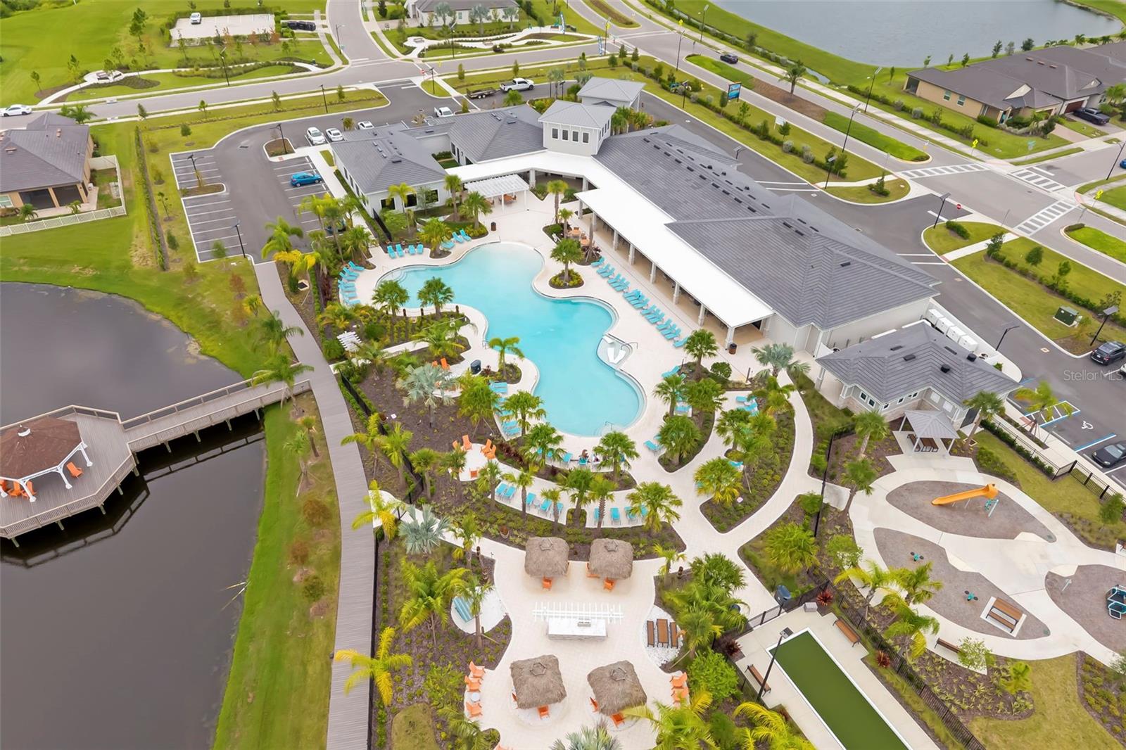 Aerial view of a community clubhouse and curving pool surrounded by palm trees, cabanas, and lounge chairs.
