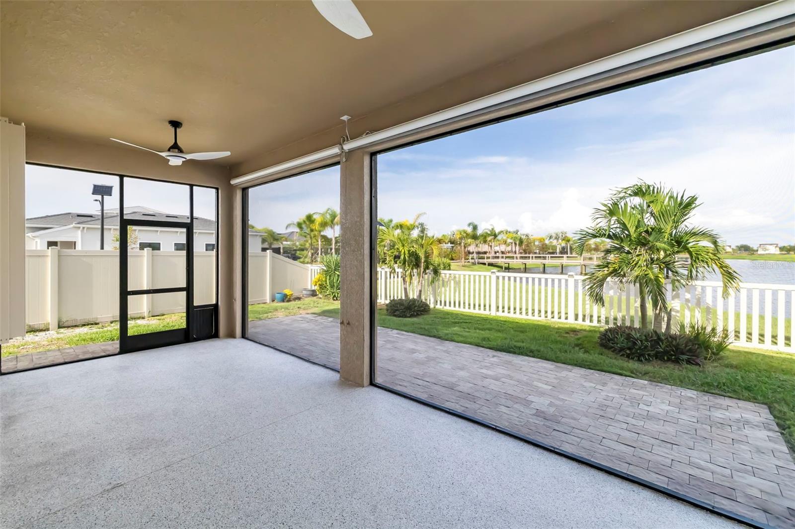 Covered lanai overlooking a fenced waterfront yard, with palm trees and a paver patio.
