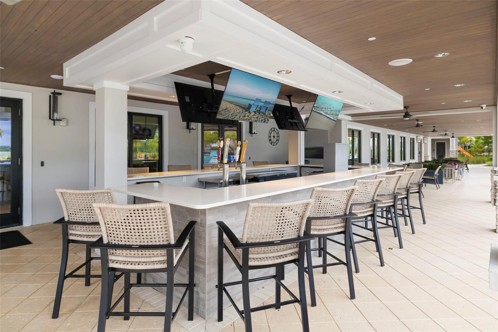 Covered outdoor bar with a long counter, woven chairs, beer taps, and suspended televisions.