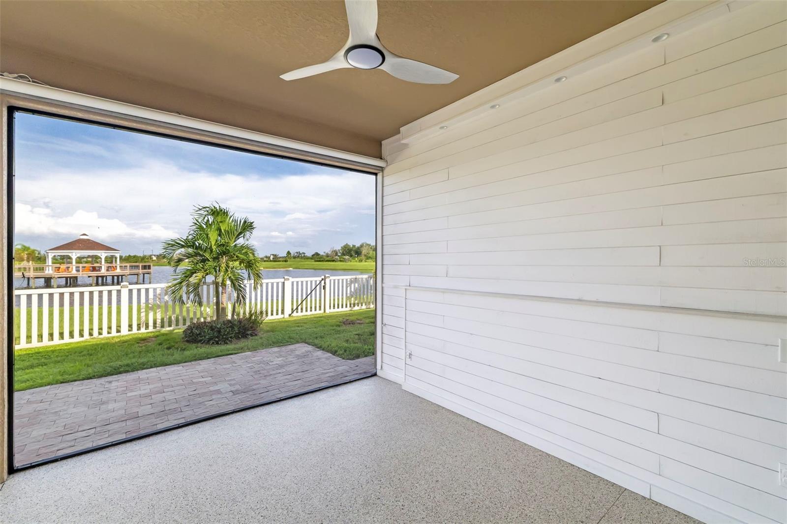 Covered patio with a ceiling fan and wide lake view, including a palm and gazebo on a dock.