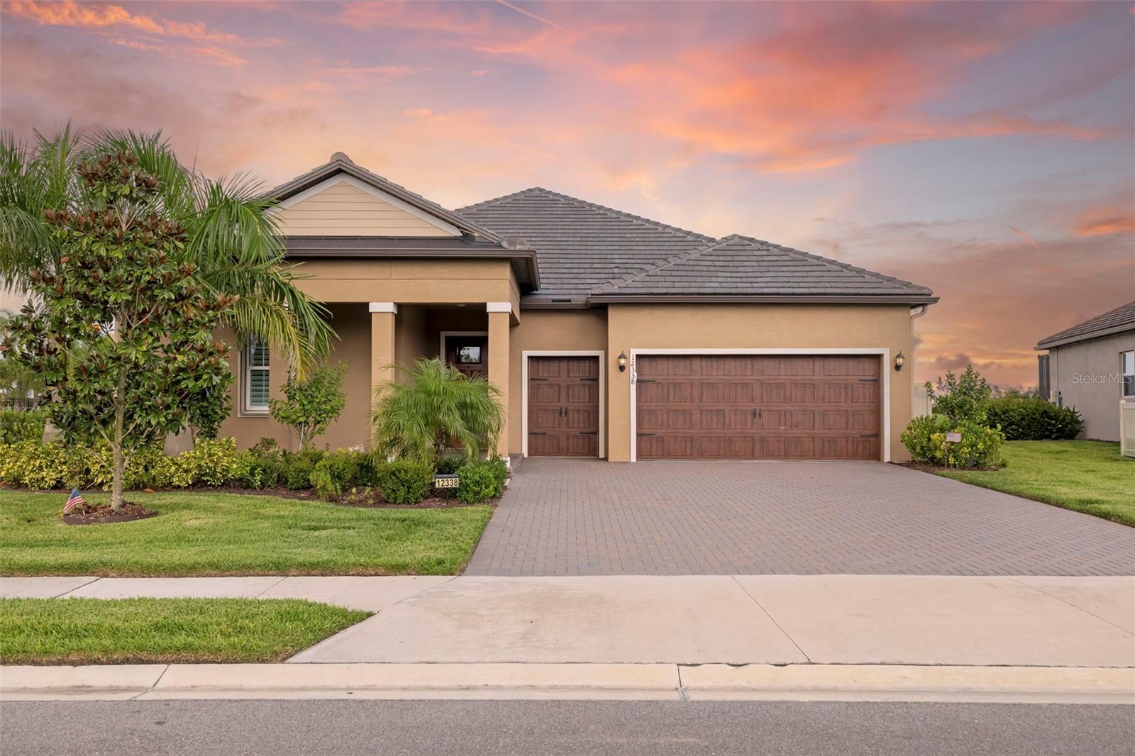 Tan single-story home with a wide wooden garage door and tropical landscaping at sunset.