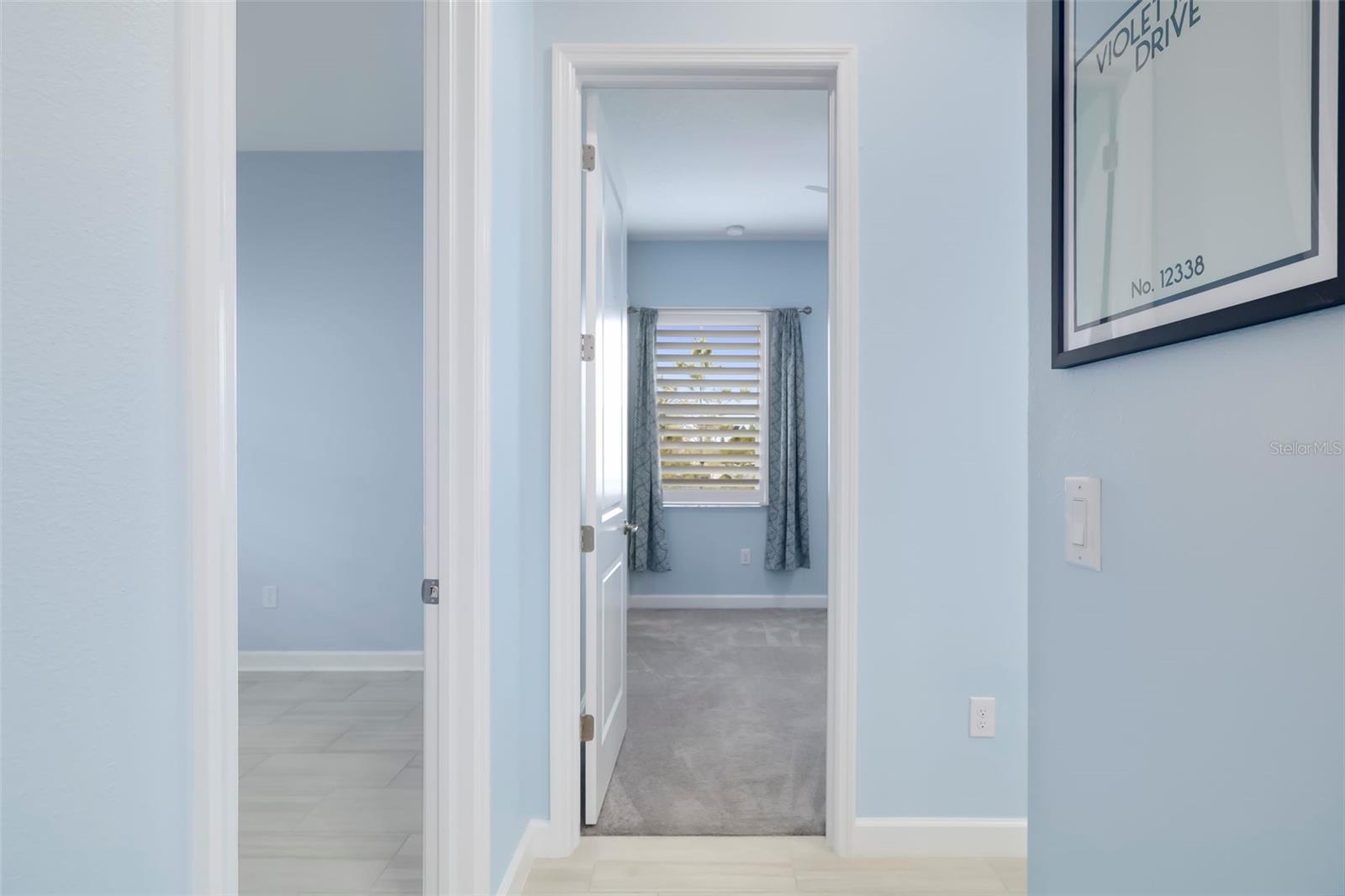 Hallway with pale blue walls and a doorway to a carpeted room with a shuttered window.