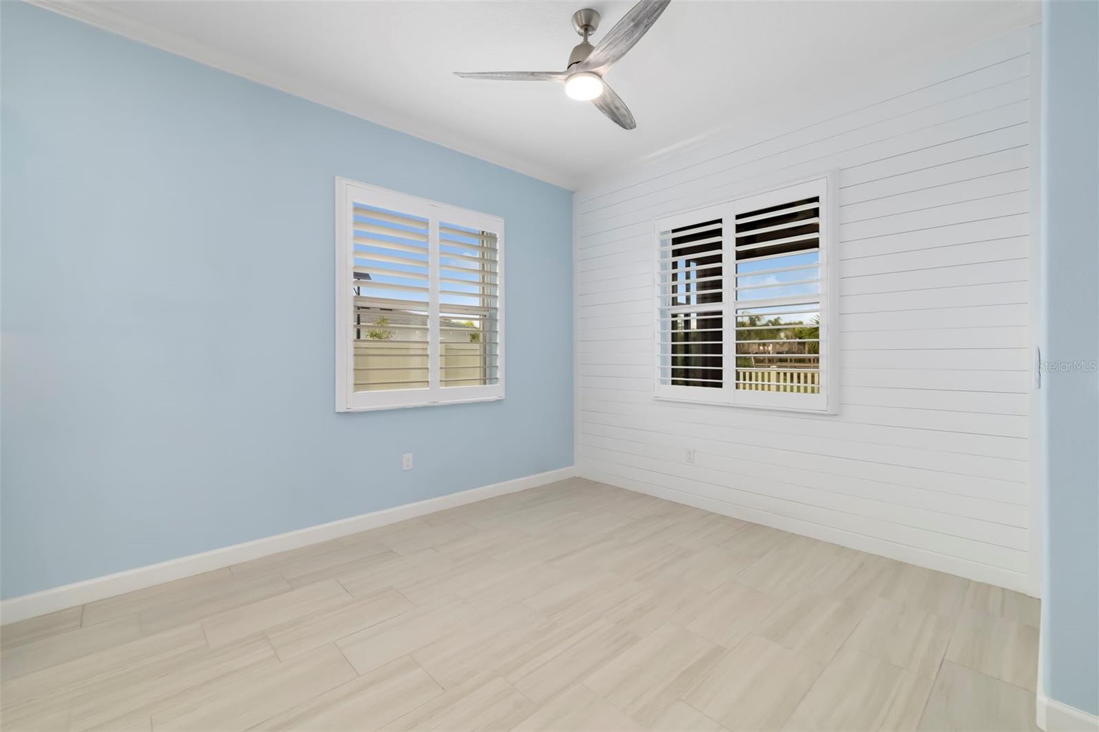 Empty room with pale blue and white shiplap walls, plantation-shuttered windows, and light tile flooring.