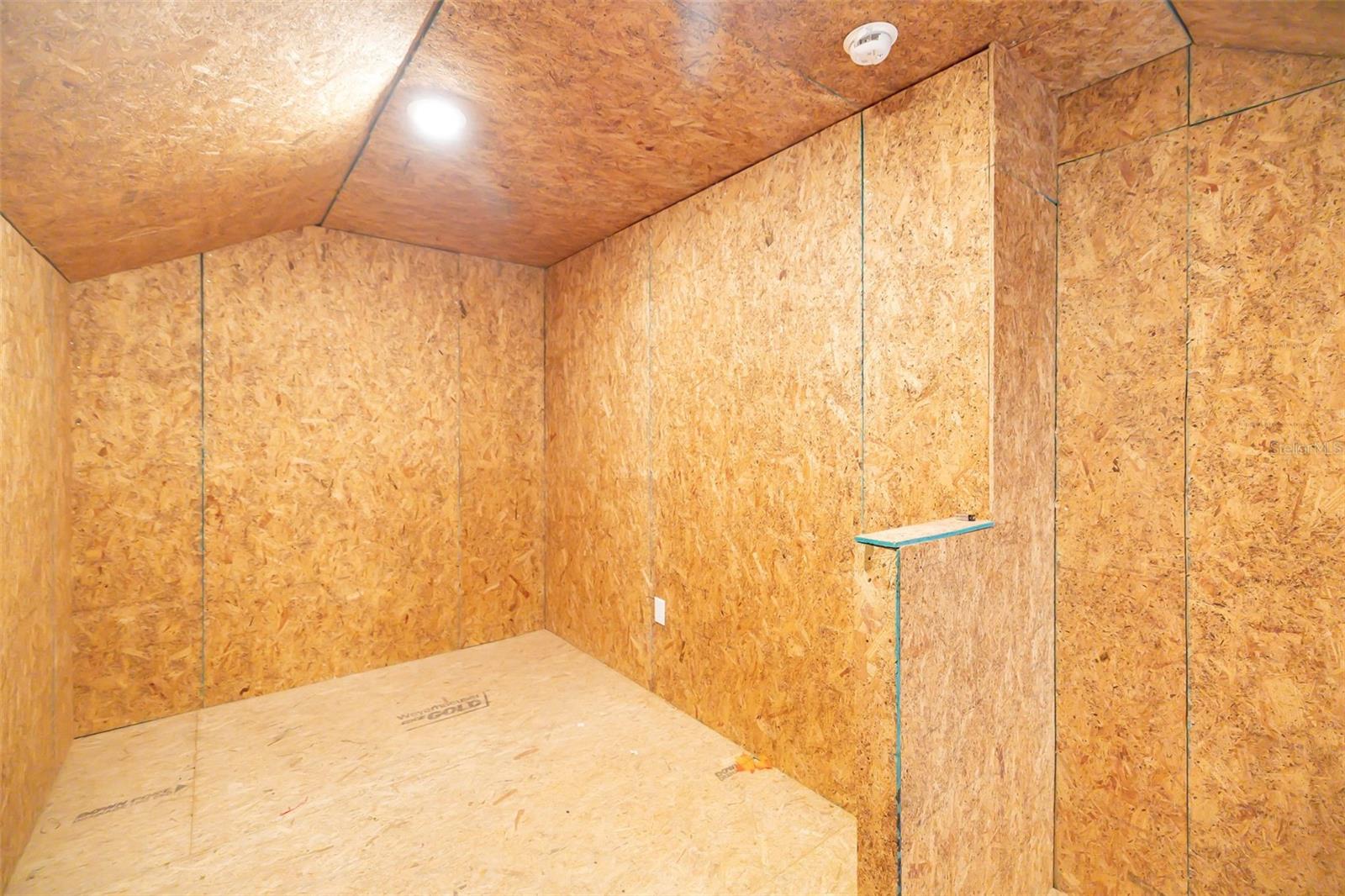 Unfinished room lined with OSB panels, with a plywood floor and narrow wall shelf.