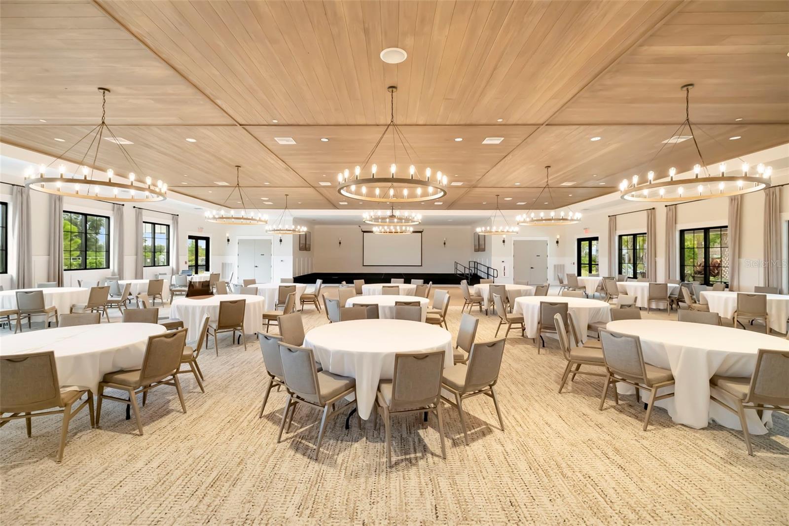 Community event room with round dining tables, circular chandeliers, and a small presentation stage.