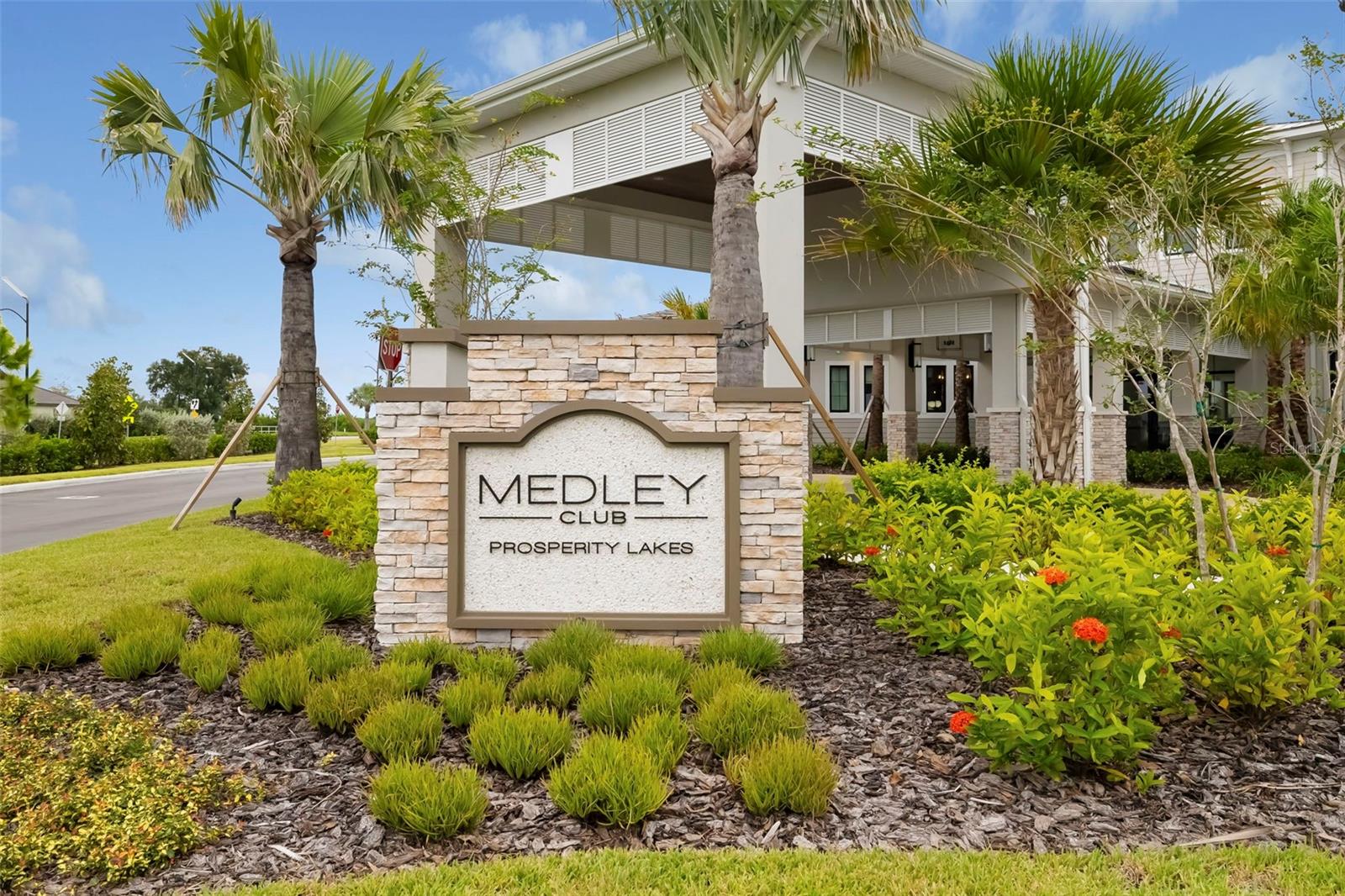 Medley Club entrance at Prosperity Lakes, marked by a stone sign amid palms and tropical landscaping.