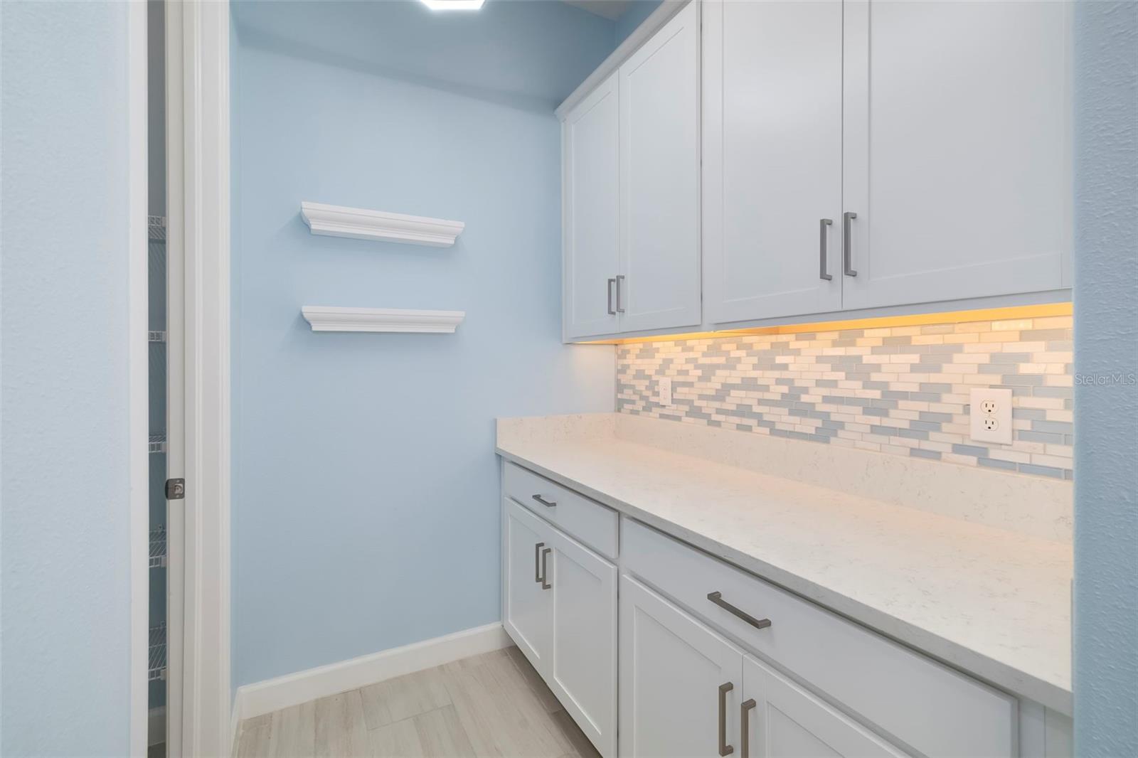 Laundry room with white cabinets, a long countertop, tile backsplash, and floating shelves.