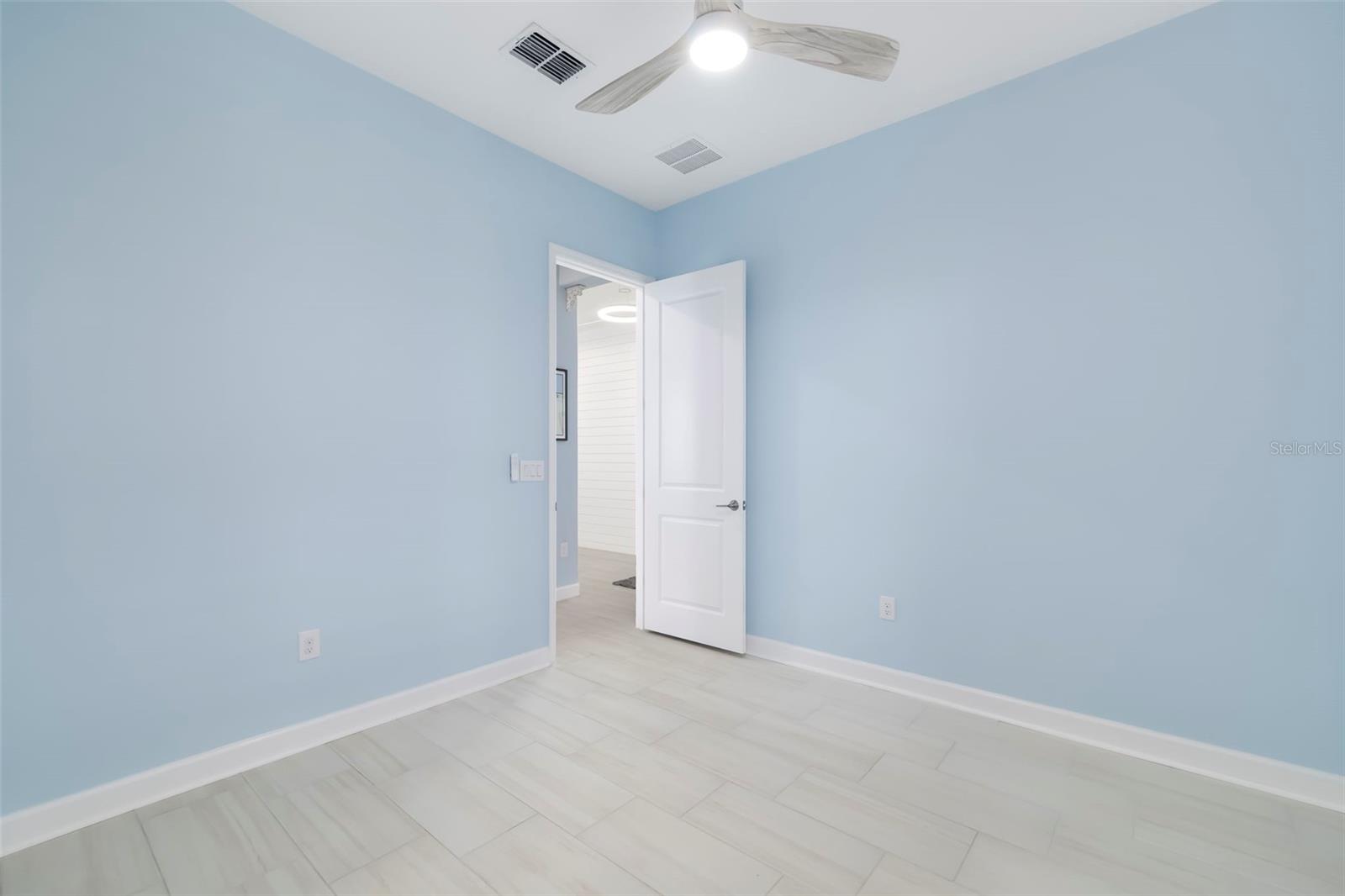 Empty pale blue room with large tile flooring, a ceiling fan, and an open door.