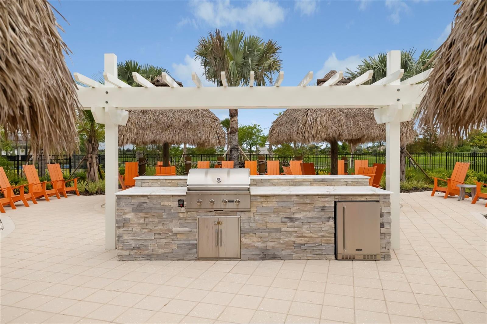 Outdoor kitchen with a stone island and stainless-steel grill beneath a pergola, surrounded by orange chairs and thatched shades.