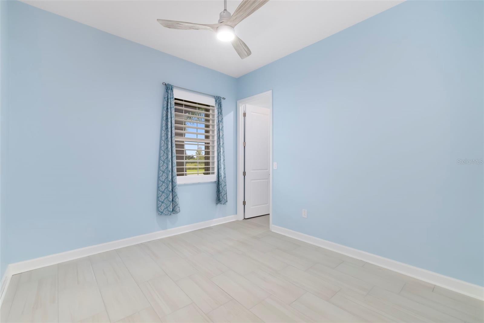 Vacant bedroom with blue walls, light tile floors, shuttered window, and ceiling fan.