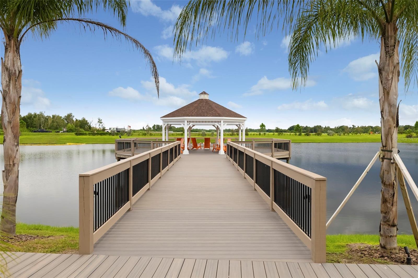 Wooden dock crosses a pond to a covered gazebo, framed by palm trees.