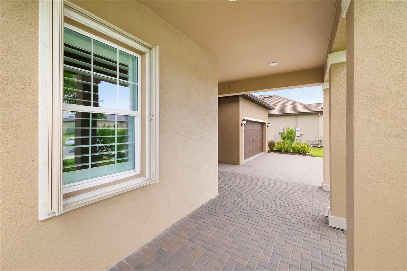Covered stucco walkway with a white-trimmed window and paver driveway leading toward a garage.