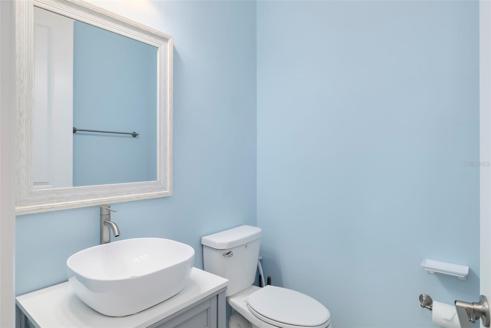 Bathroom with pale blue walls, a vessel sink, toilet, and large white-framed mirror.