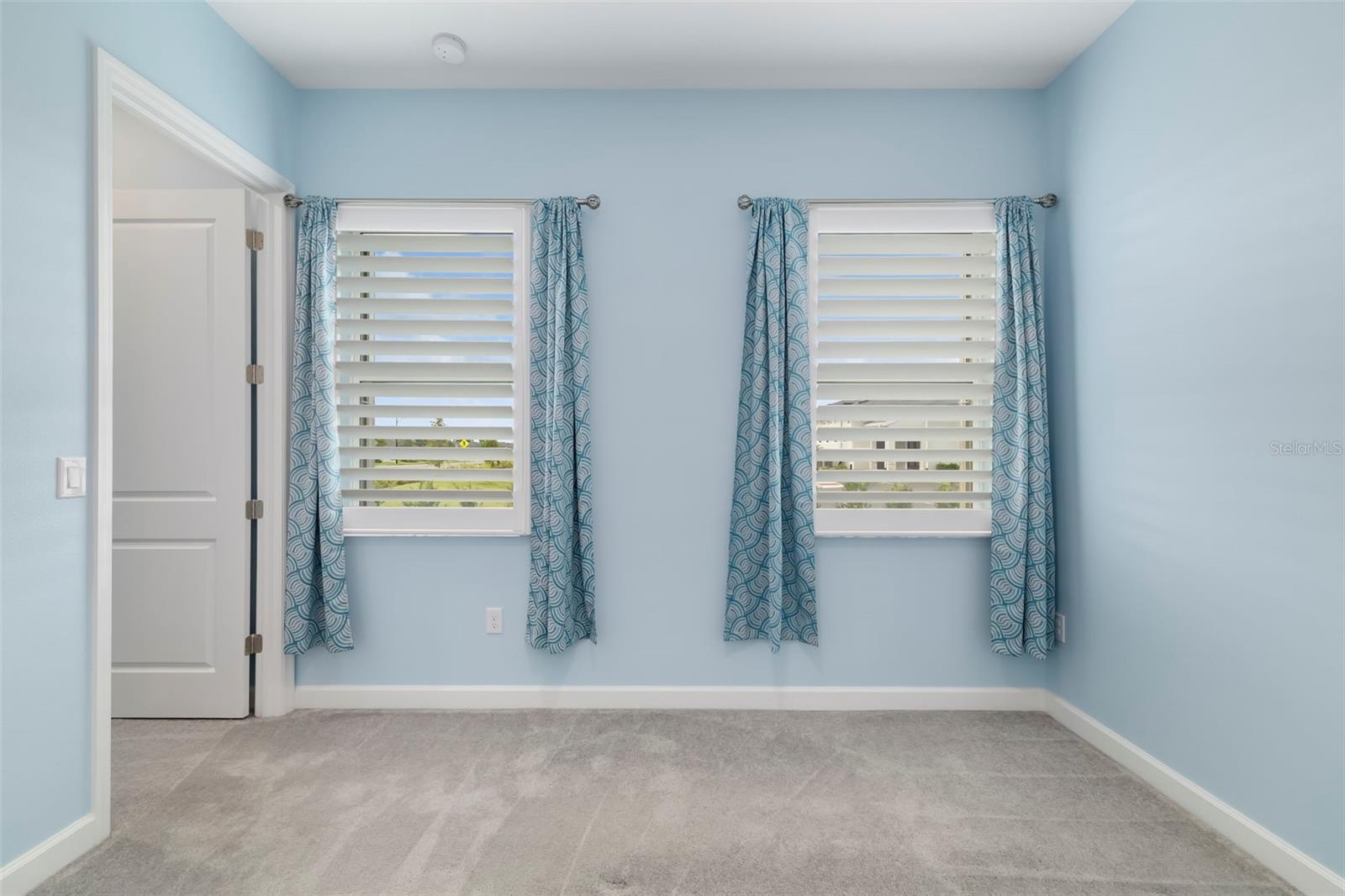 Empty blue bedroom with gray carpet and two curtained windows.