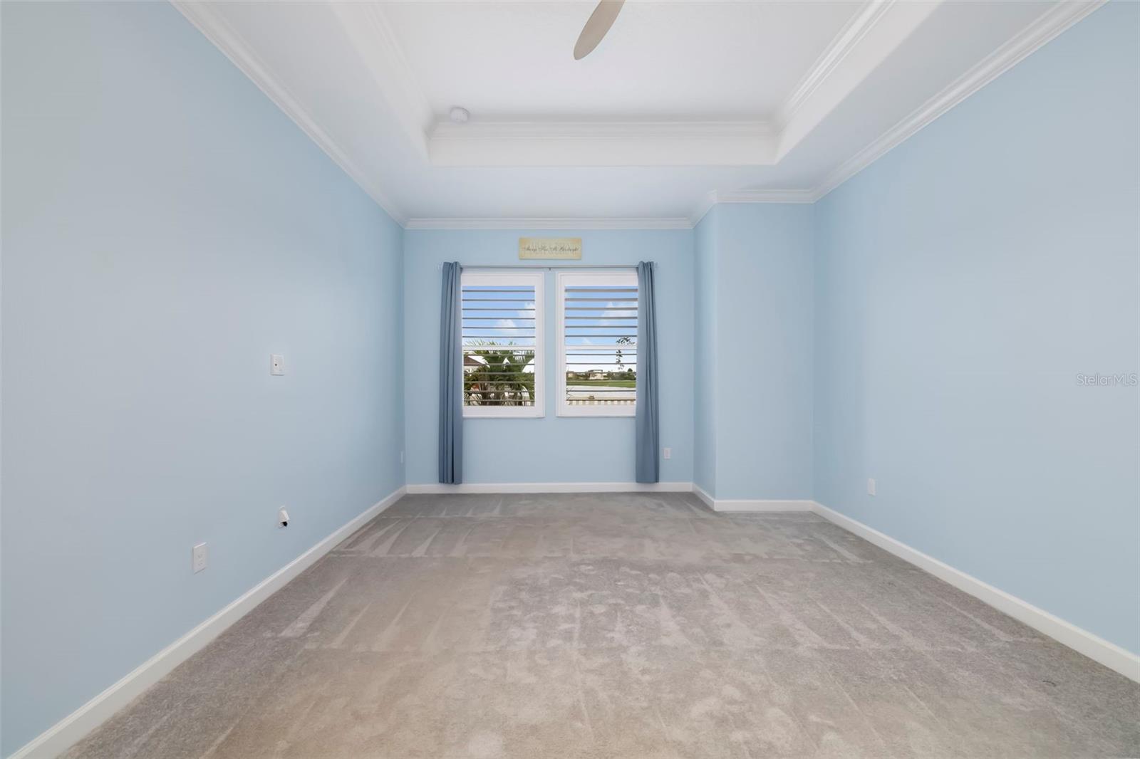 Empty blue bedroom with a tray ceiling, gray carpet, and windows overlooking water.
