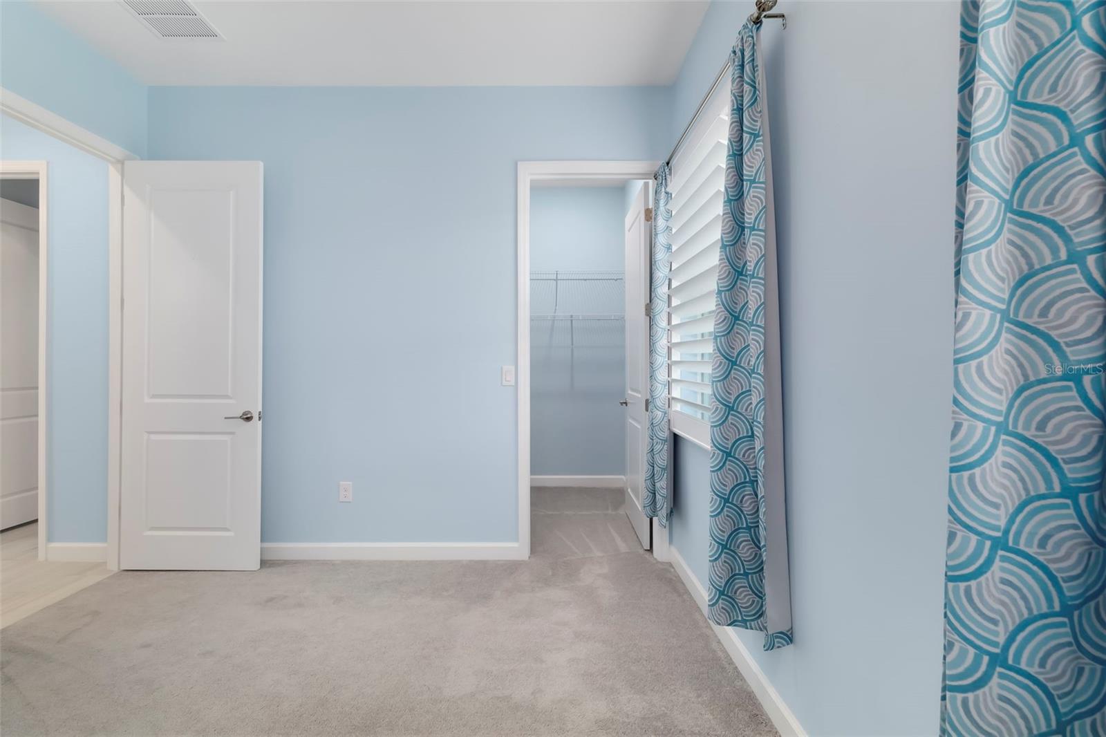 Empty blue bedroom with beige carpet, wire closet shelving, and teal patterned curtains.