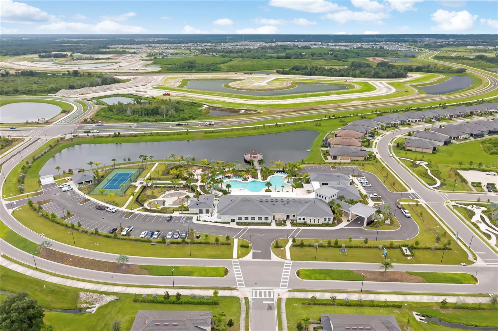 Aerial view of a community clubhouse, pool, tennis court, pond, and surrounding homes.