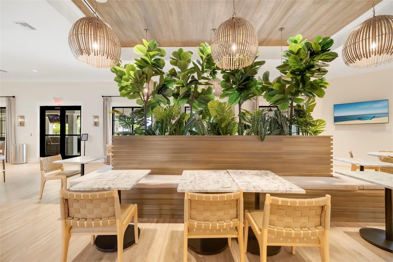 Communal dining area with marble-top tables, woven chairs, a wood banquette, and tall indoor plants.