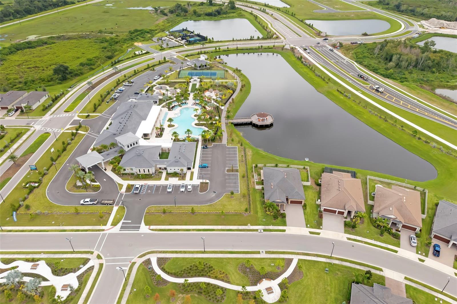 Aerial view of a community clubhouse and pool beside a large lake, with tennis courts and nearby homes.