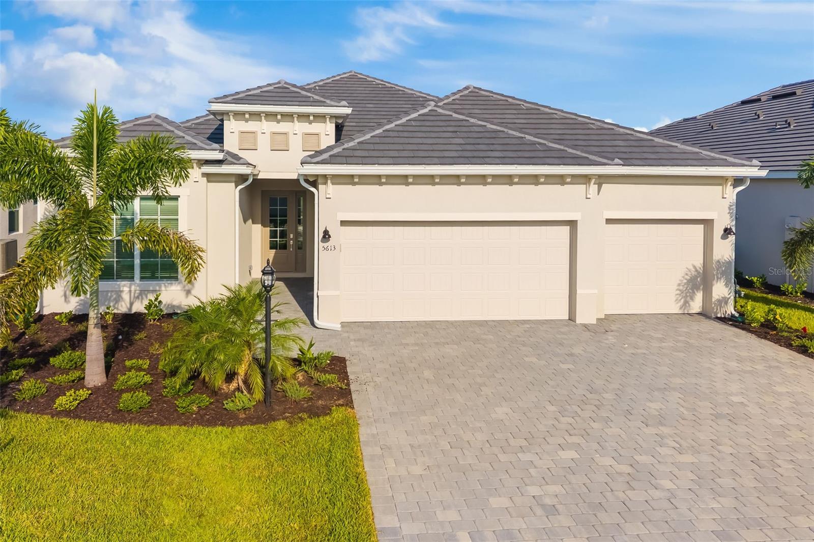 Beige home with a tiled roof, two-car garage, paver driveway, and palm landscaping.