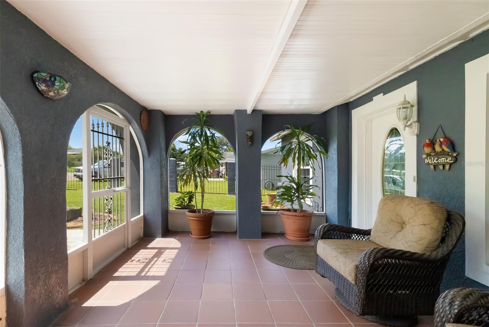 Covered porch with arched openings, pink tile floor, potted plants, and wicker seating.