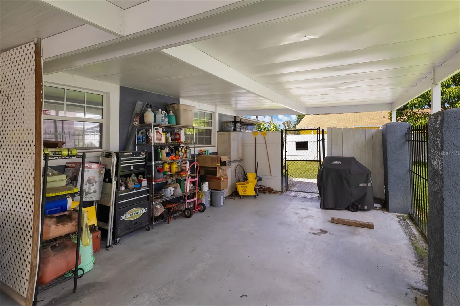 Covered carport with storage shelves and tool chests, leading to a gated grassy area.