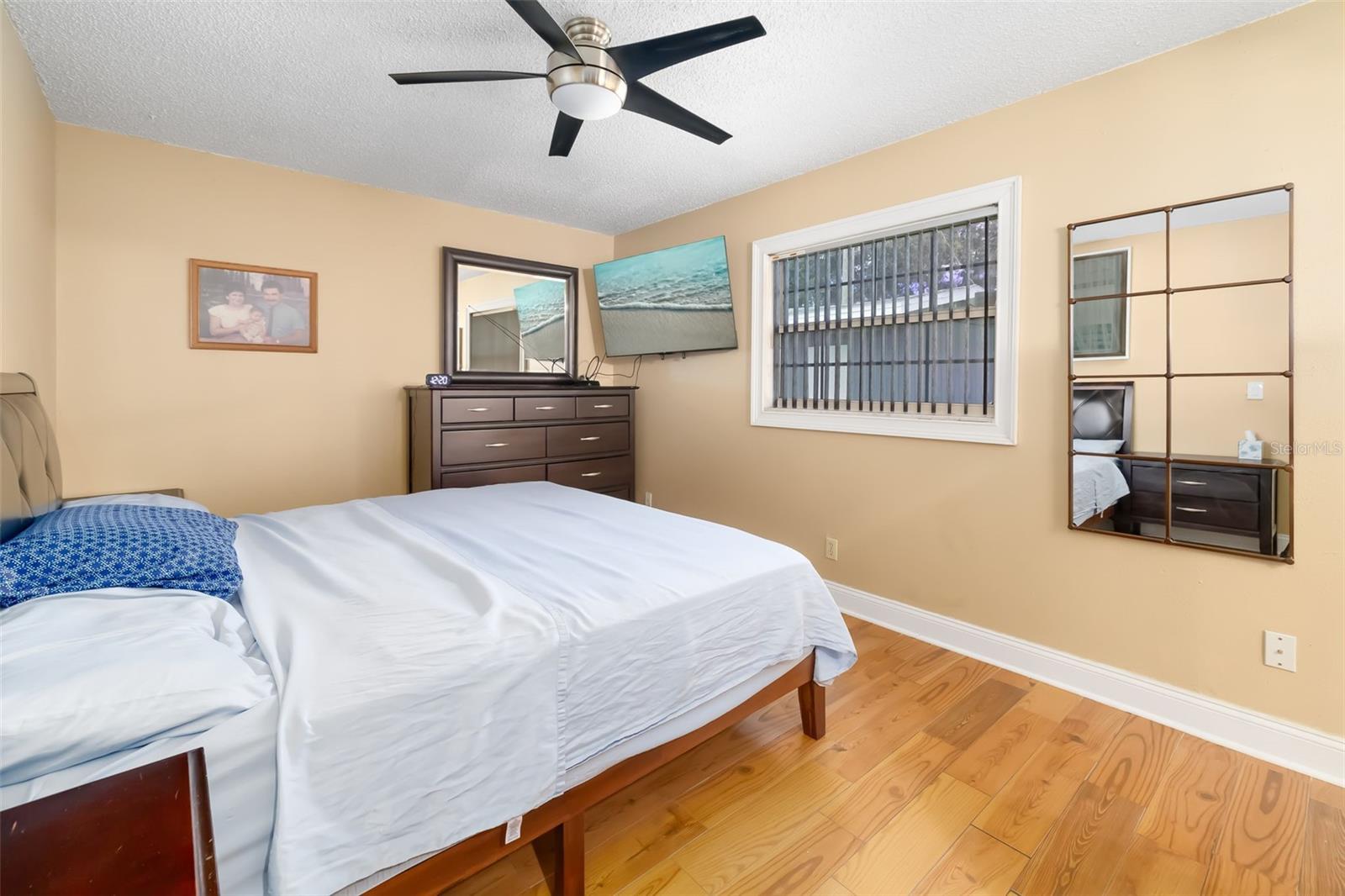 Bedroom with a white-covered bed, hardwood floors, and a dark dresser beneath a mirror.