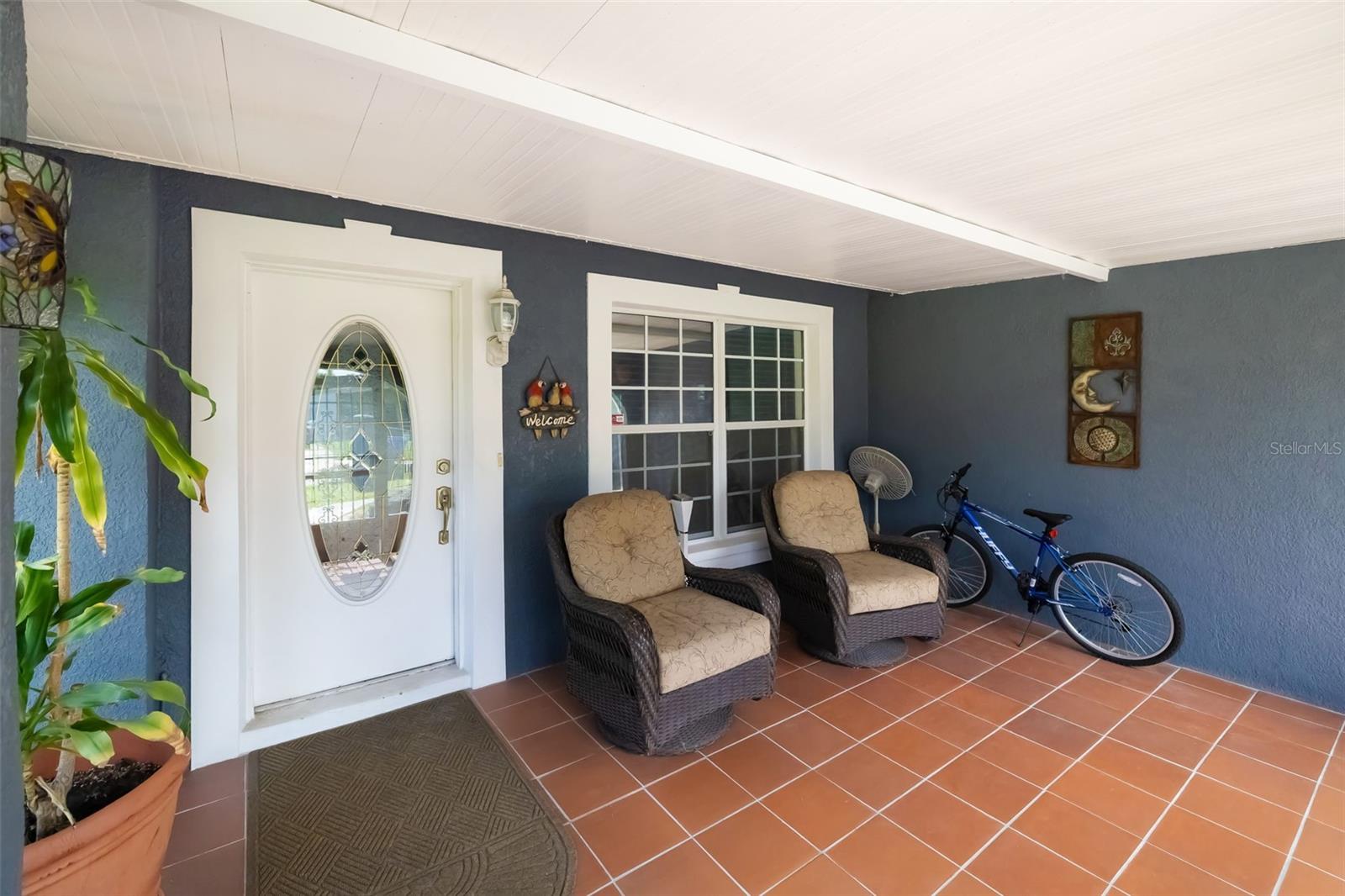 Covered front porch with oval-glass entry door, two wicker chairs, terracotta tile, and a bicycle.