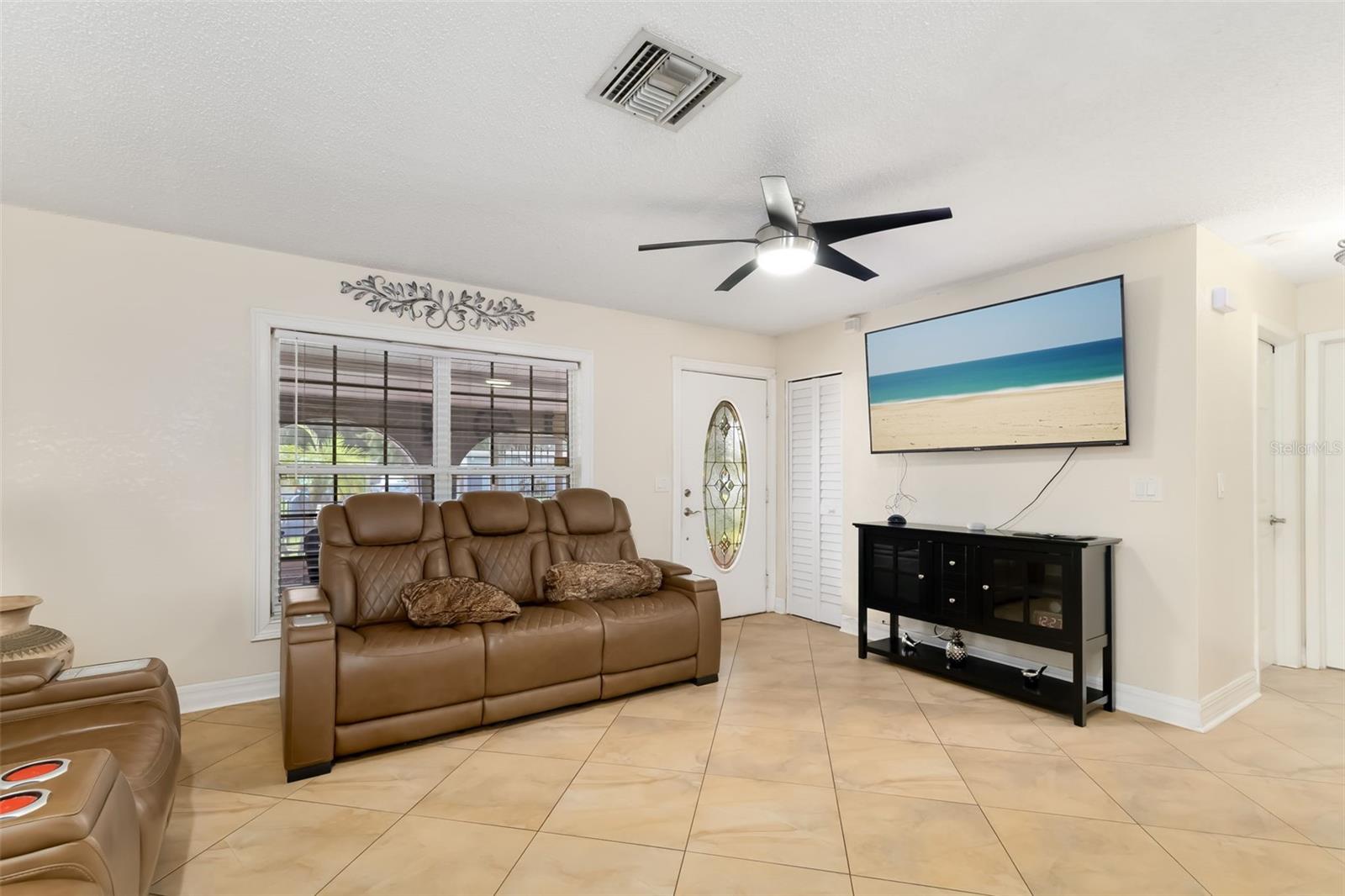 Living room with tan leather sofas, beige tile floors, and a wall-mounted television showing a beach.