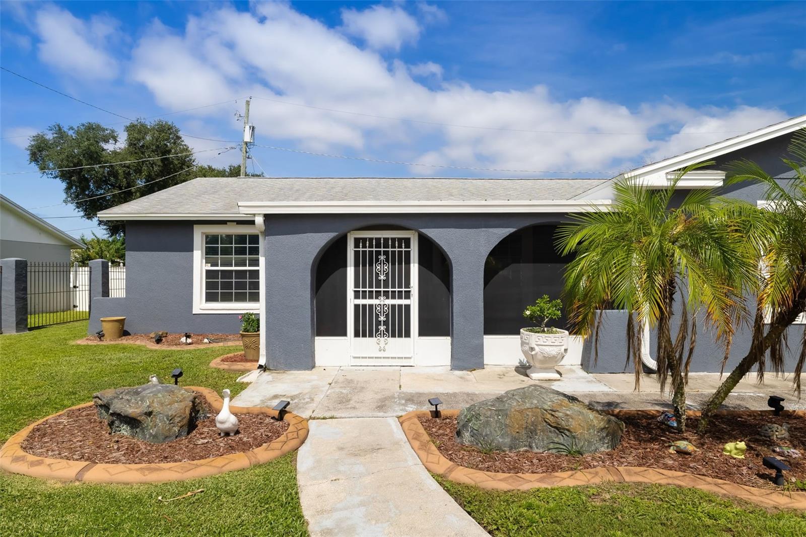 Gray stucco home with arched porch openings, white security door, palms, and a landscaped front yard.