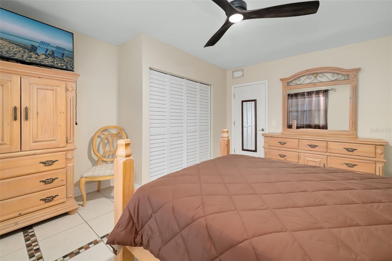 Furnished bedroom with a brown quilted bed, light wood dressers, and white louvered closet doors.