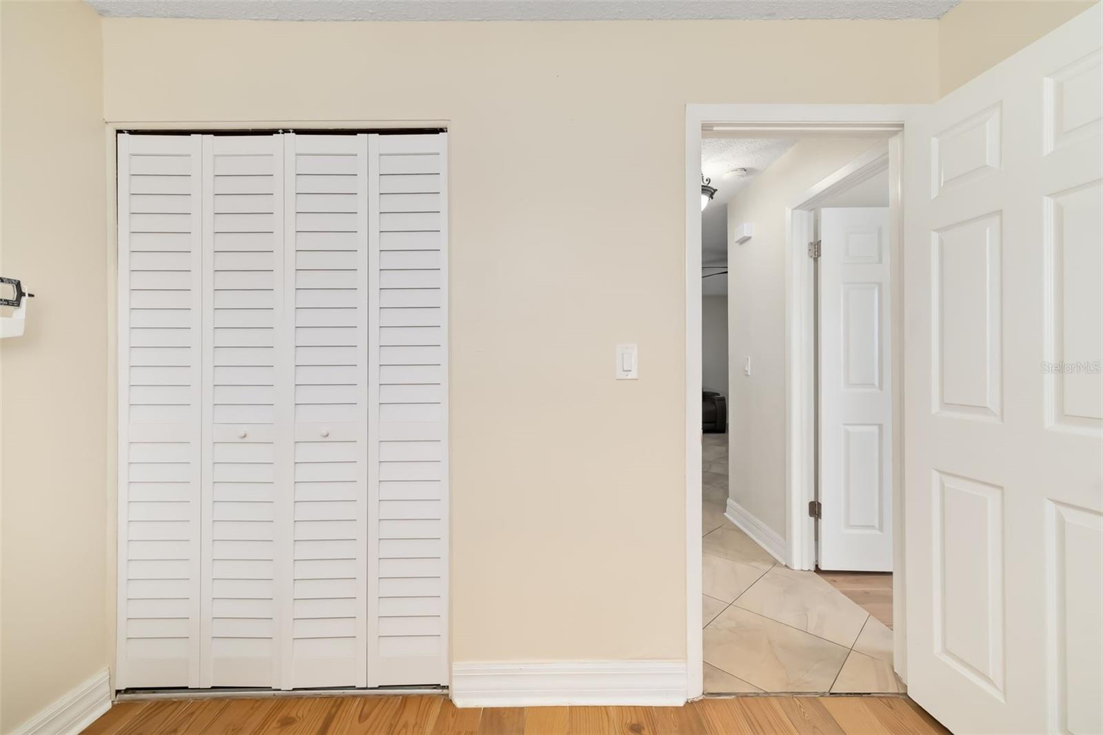 Room with white louvered closet doors, wood flooring, and an open doorway to a tiled hall.