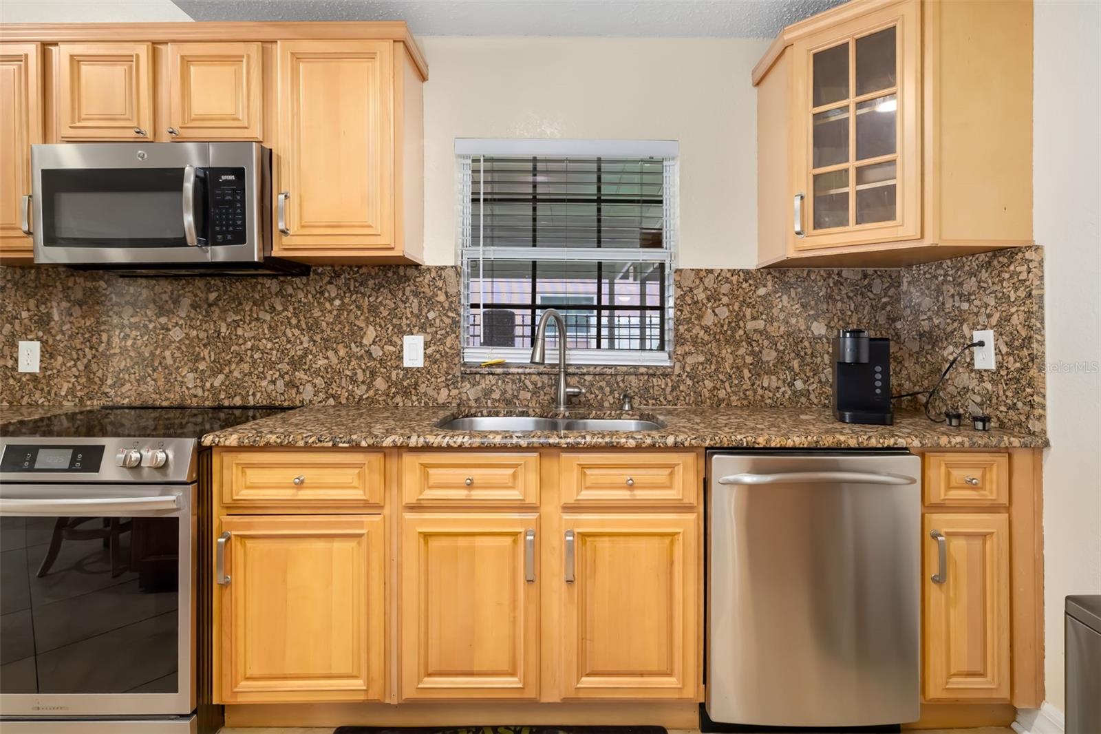Kitchen with wood cabinets, patterned granite counters, stainless appliances, and a double sink beneath a window.