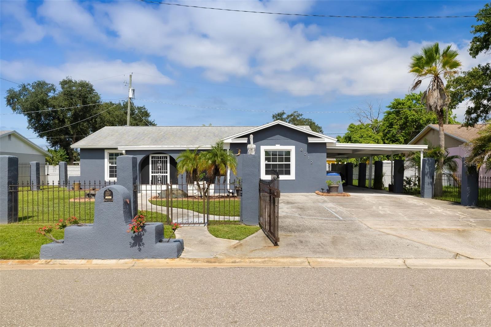 Blue-gray single-story home with a gated front yard, palm trees, and a covered carport.