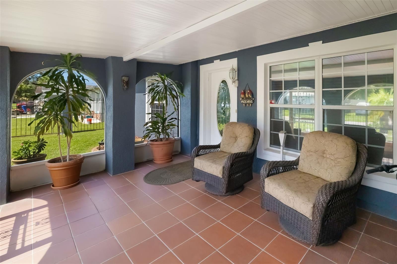 Covered front porch with arched openings, potted plants, and two wicker chairs.